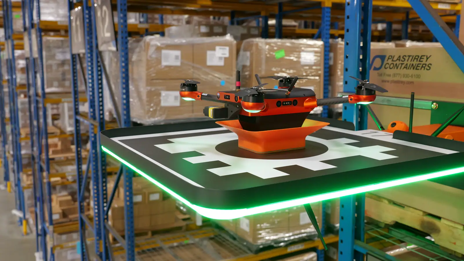 Corvus One autonomous warehouse inventory drone docked on its rack-mounted landing pad, the pad edge lit green, in a distribution center aisle