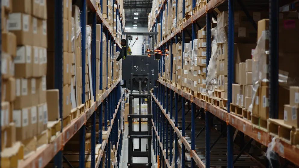 Corvus One autonomous warehouse inventory drone flying at the upper rack levels of a long aisle at GNC, alongside an order picker raised to the same height