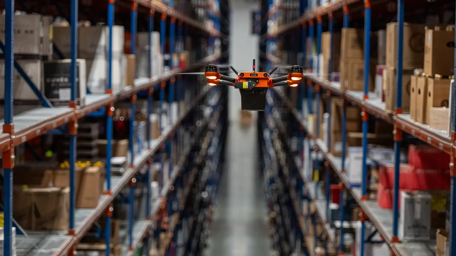 Corvus One autonomous inventory drone flying down a long aisle between tall shelving racks in a distribution center